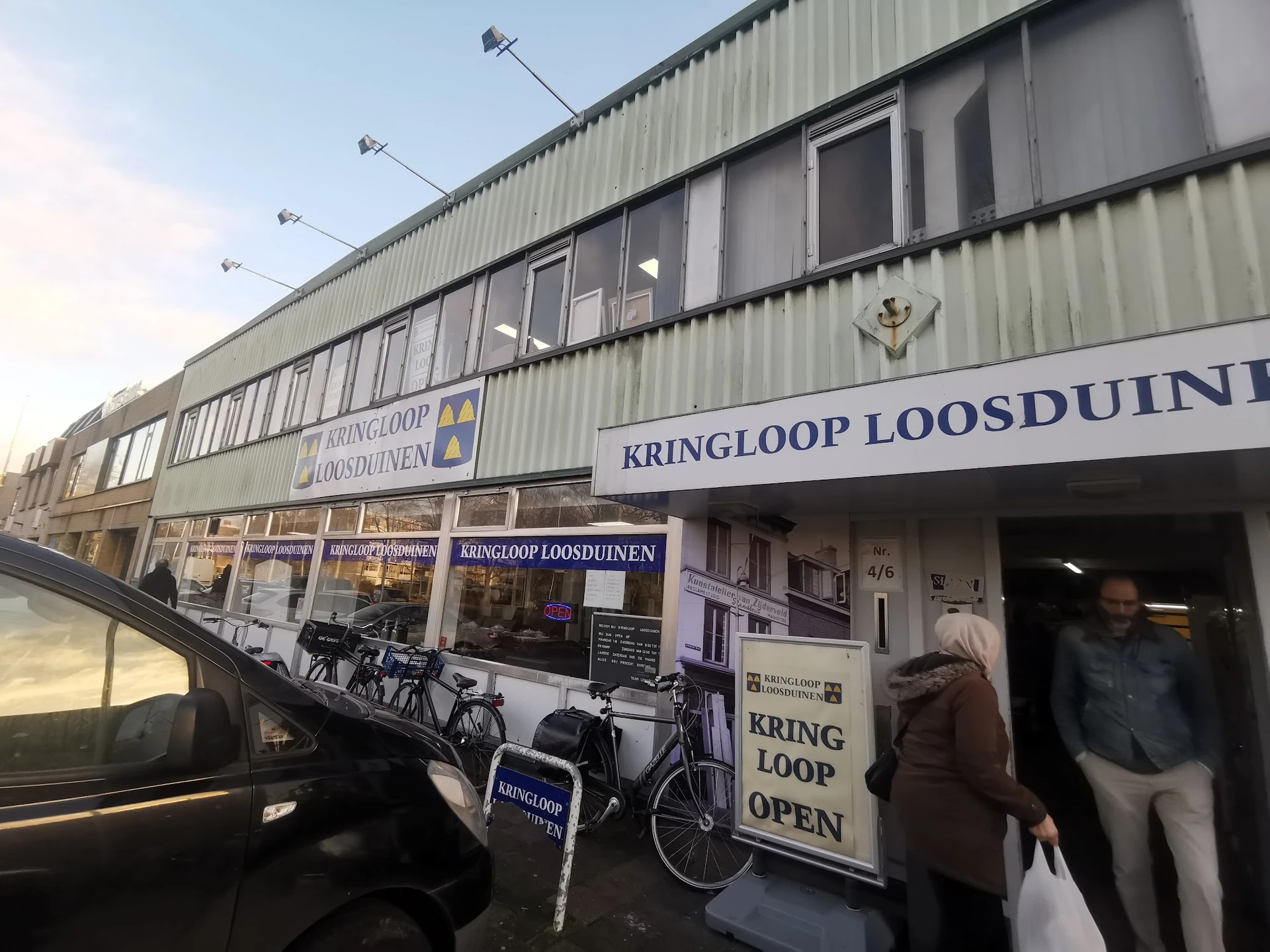 Voorgevel van kringloopwinkel met grote ramen, winkelnaam op gevel, ingang met mensen, fietsen buiten