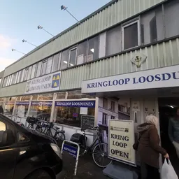 Voorgevel van kringloopwinkel met grote ramen, winkelnaam op gevel, ingang met mensen, fietsen buiten