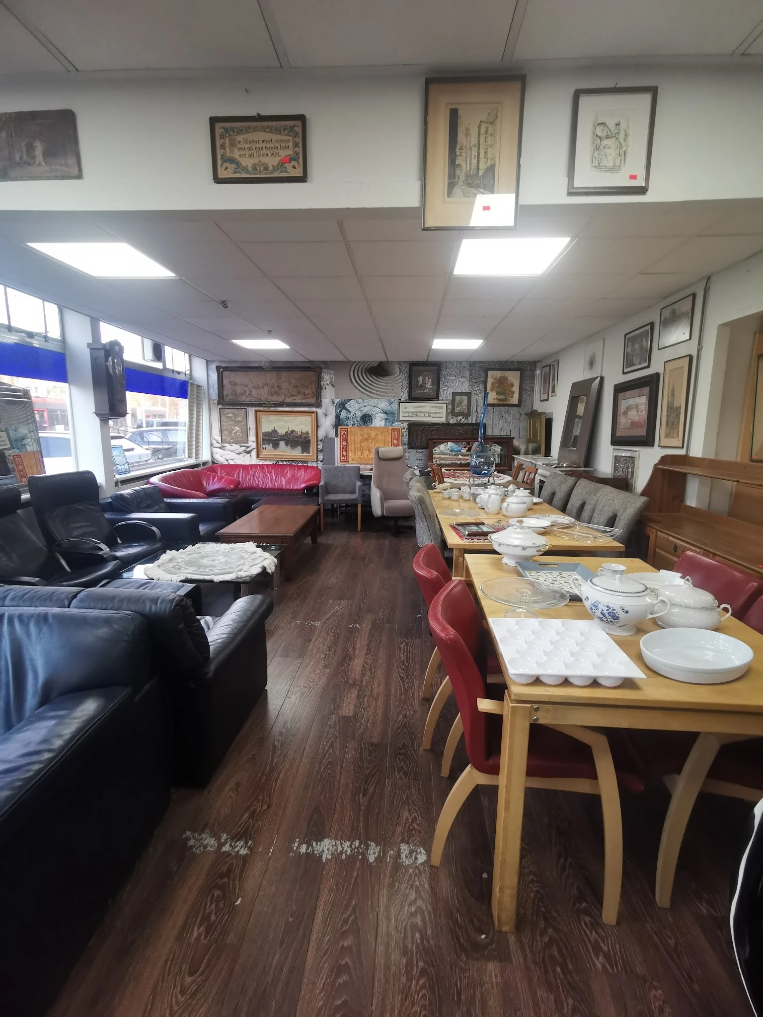 Interieur van kringloopwinkel met zithoek met zwarte banken, eettafels met borden en bestek, en aan muur hangend aanbod van s