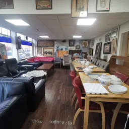 Interieur van kringloopwinkel met zithoek met zwarte banken, eettafels met borden en bestek, en aan muur hangend aanbod van s