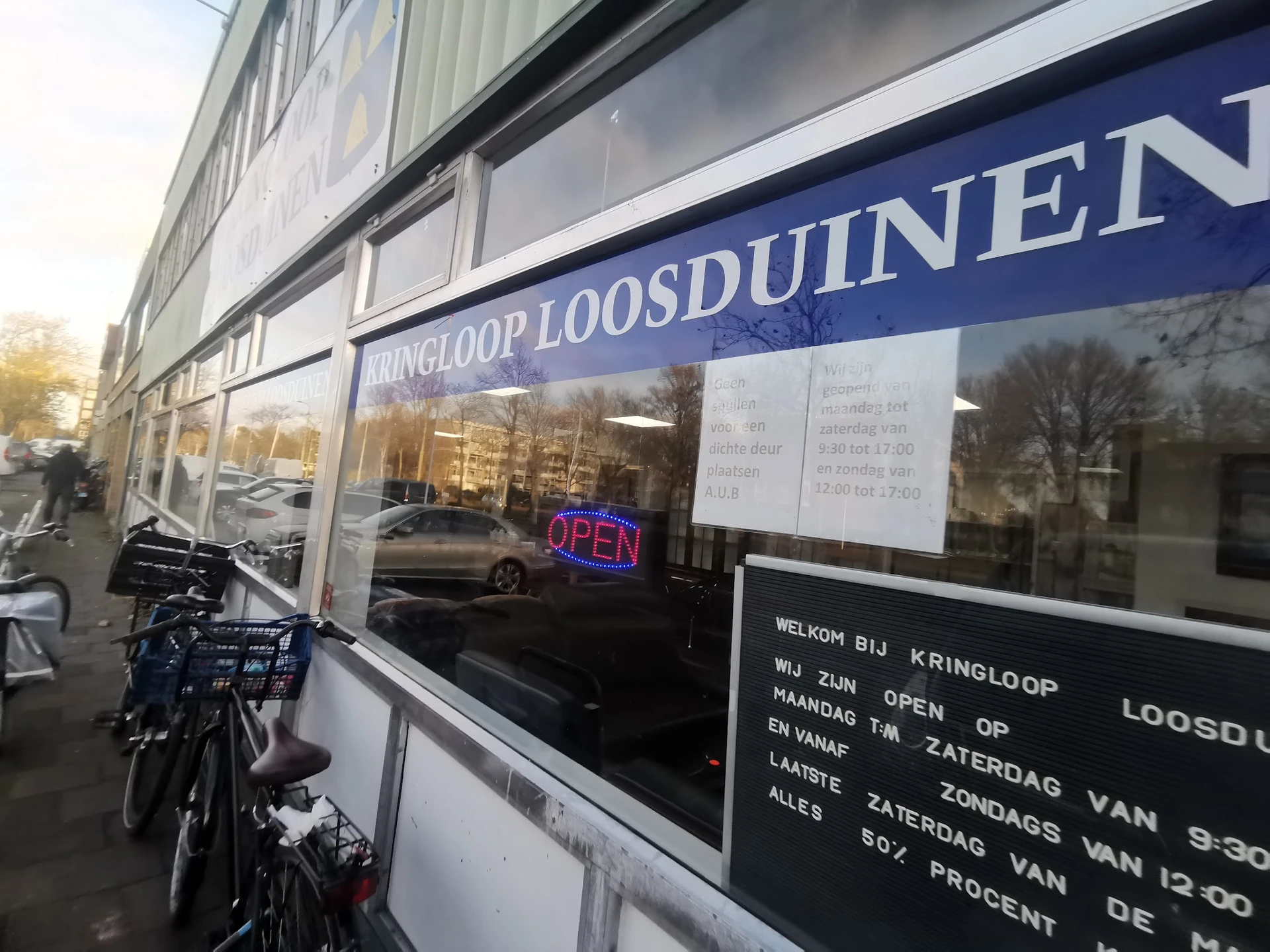 Gevel van kringloopwinkel met blauwe luifel, winkelnaam op raam en openingstijden zichtbaar