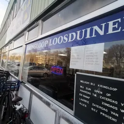 Gevel van kringloopwinkel met blauwe luifel, winkelnaam op raam en openingstijden zichtbaar