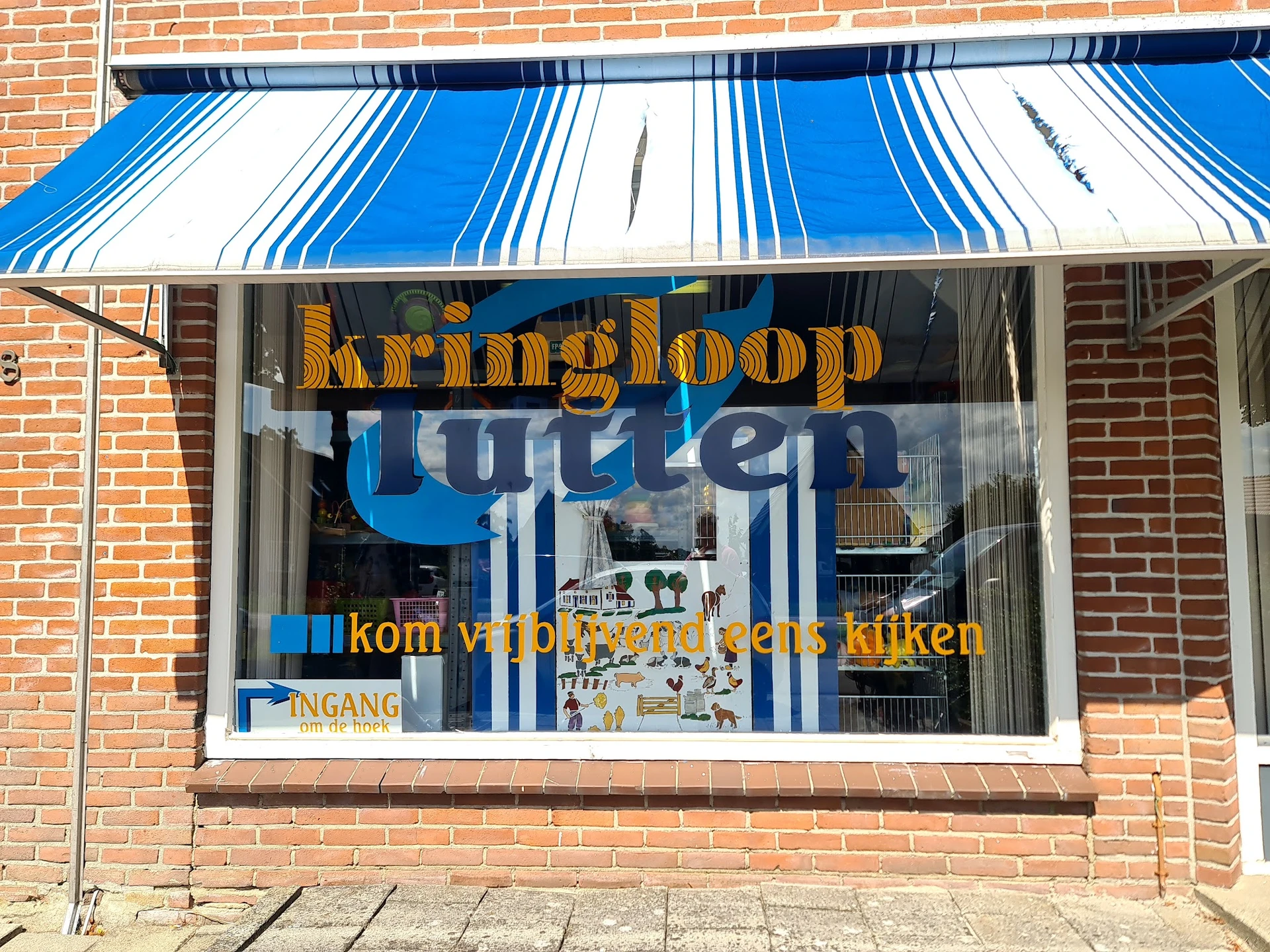Gevel van kringloopwinkel met blauwe luifel, grote ramen en gele/blauwe letters op het glas