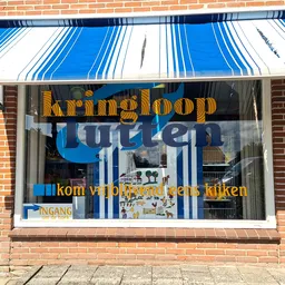 Gevel van kringloopwinkel met blauwe luifel, grote ramen en gele/blauwe letters op het glas