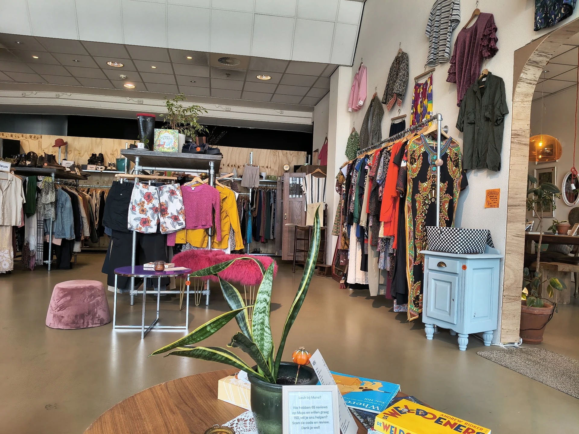Winkelvloer van kringloopwinkel met kledingrekken, stellingen met tweedehands kleding en een plant op een tafel op de voorgro