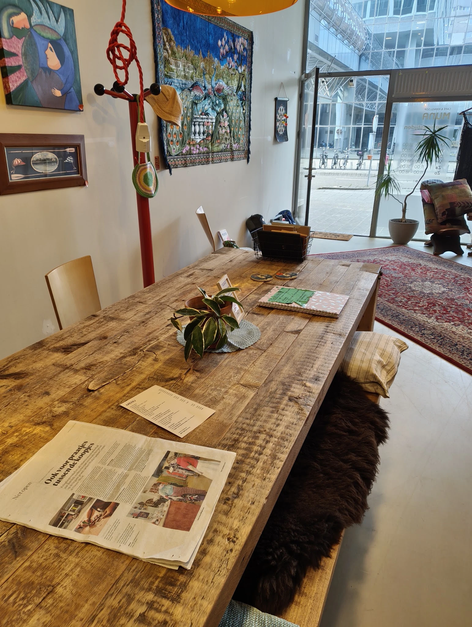 Houten tafel met stoelen, plant en papieren in een kringloopwinkel met zicht op de winkelruit en straat