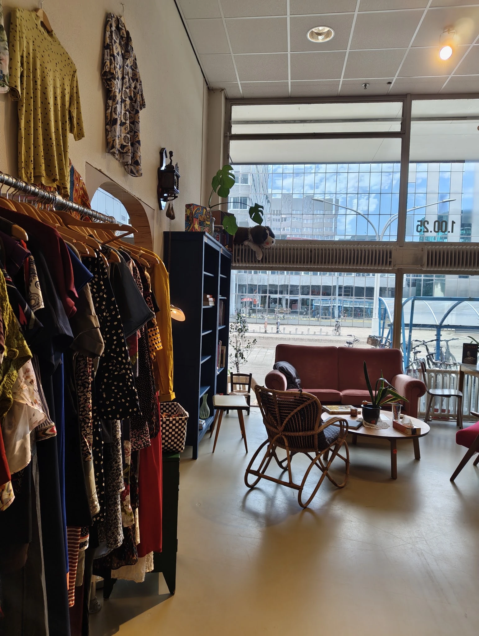 Winkelinterieur met kledingrekken links, zithoek met rode bank en stoelen in het midden, grote ramen met uitzicht op straat