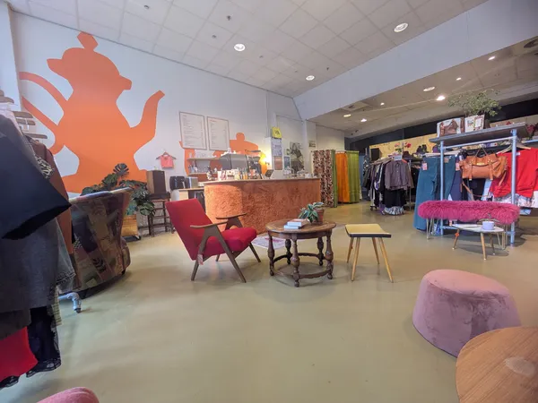 Winkelvloer van kringloopwinkel met rode fauteuils, tafels, kledingrekken en grote oranje decoratie aan de muur
