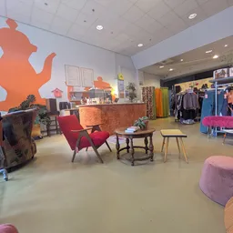 Winkelvloer van kringloopwinkel met rode fauteuils, tafels, kledingrekken en grote oranje decoratie aan de muur
