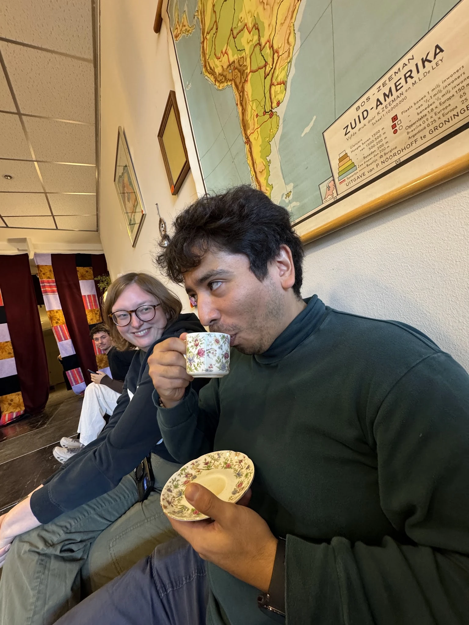 Man drinkt uit een mok met bloemenpatroon terwijl een vrouw in de achtergrond toekijkt in een kringloopwinkel.