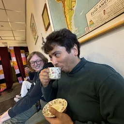 Man drinkt uit een mok met bloemenpatroon terwijl een vrouw in de achtergrond toekijkt in een kringloopwinkel.