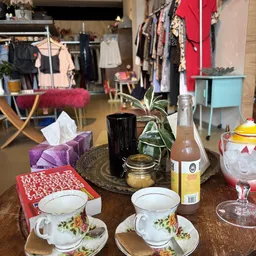 Tafelset met glazen, fles, plant en serviesgoed in een kringloopwinkel met kledingrekken op achtergrond