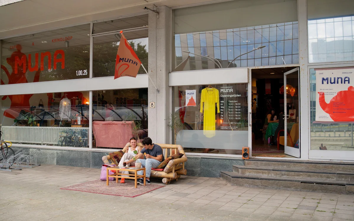 Winkelgevel met etalages en twee personen zittend op een houten bank op het terras ervoor