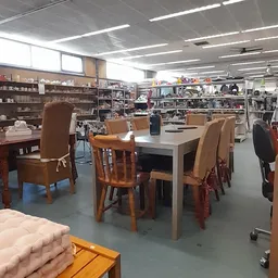 Winkelvloer met eettafelset in het midden, stellingen en kasten vol spullen op de achtergrond, heldere verlichting