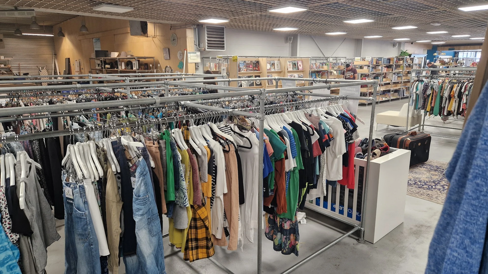 Rekken met tweedehands kleding op hangers in kringloopwinkel, op achtergrond boeken in kasten