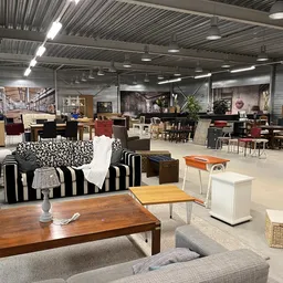 Ruime winkelhal met diverse meubels en huishoudartikelen uitgestald, met gestreepte bank, houten tafels en stoelen
