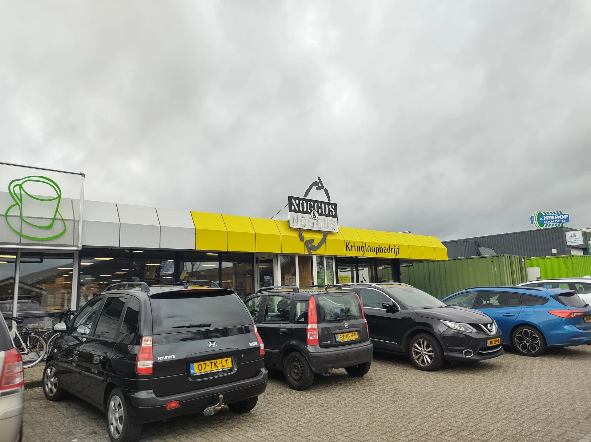Voorkant van kringloopwinkel Noggus met geel afdak, parkeerplaats met auto's en bewolkte lucht