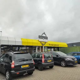 Voorkant van kringloopwinkel Noggus met geel afdak, parkeerplaats met auto's en bewolkte lucht