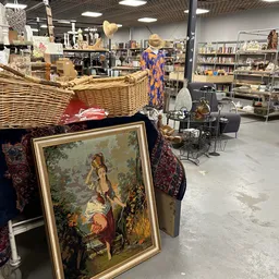 Diverse tweedehands spullen uitgestald in een kringloopwinkel, waaronder een geschilderd portret, rieten mand, kleding en hui