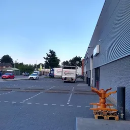 Parkeerterrein met bestelwagen en hoek van bakstenen gebouw, bomen op achtergrond