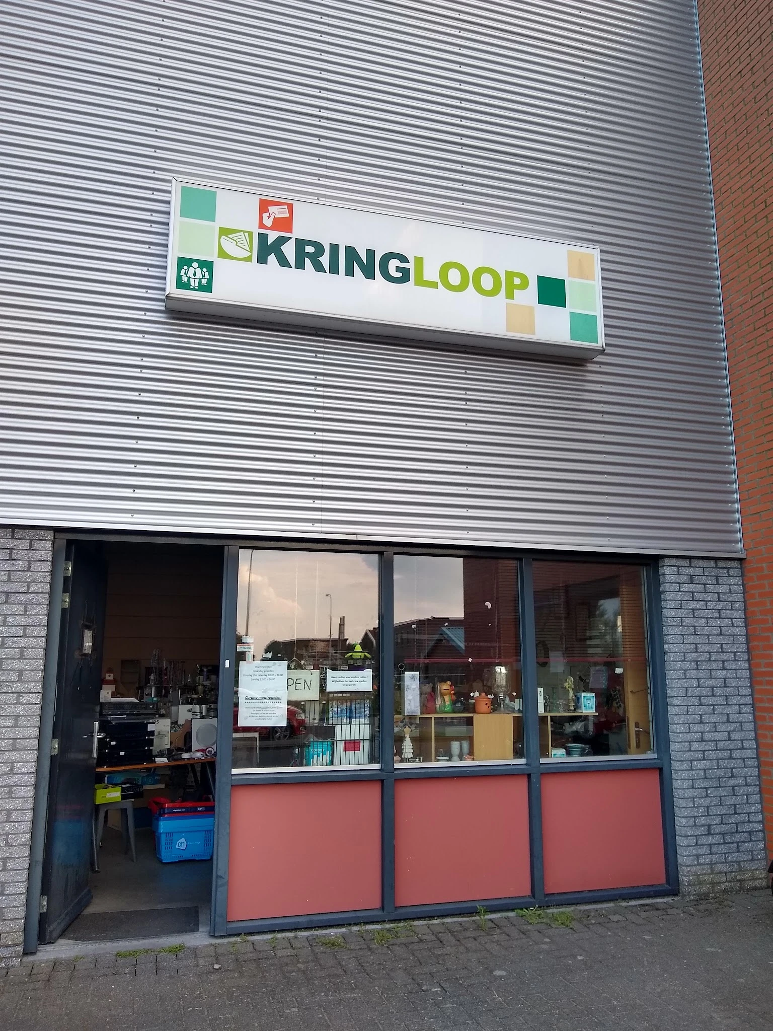 Gevel van kringloopwinkel met witte golfplaten, groene KRINGLOOP-logo, bakstenen zijkant en grote ramen met winkeluitstalling