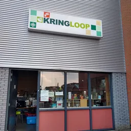 Gevel van kringloopwinkel met witte golfplaten, groene KRINGLOOP-logo, bakstenen zijkant en grote ramen met winkeluitstalling