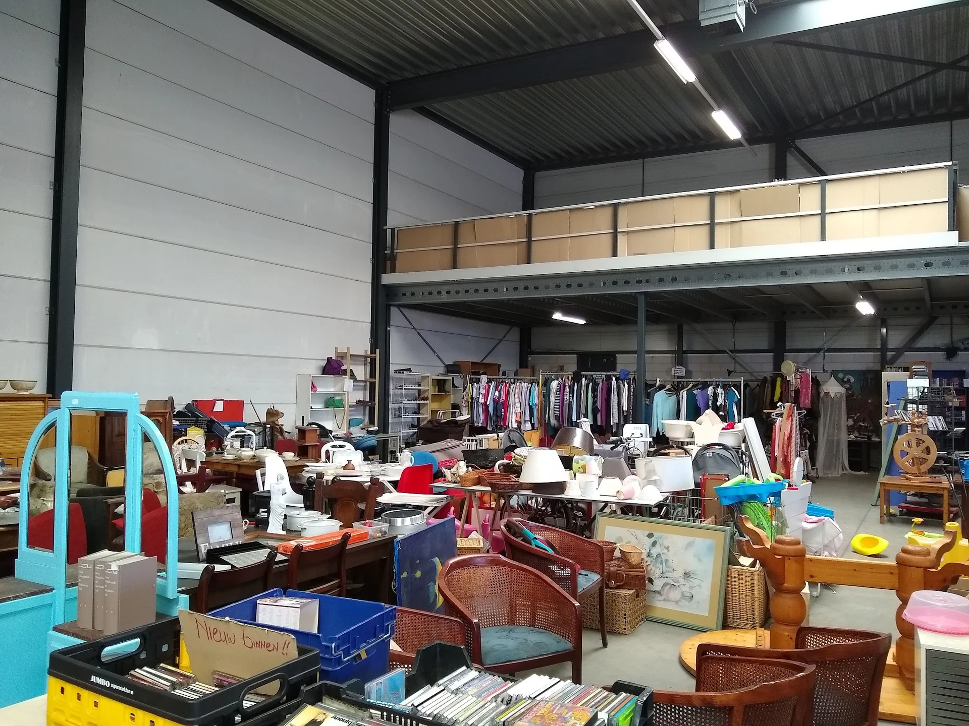 Overzicht van winkelvloer met diverse tweedehands spullen, kleding op rekken en opgestapelde huishoudelijke artikelen