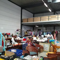 Overzicht van winkelvloer met diverse tweedehands spullen, kleding op rekken en opgestapelde huishoudelijke artikelen