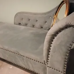 Grijze fauteuil met goudkleurige houten armleuningen en zilveren siernagels, close-up van zijkant