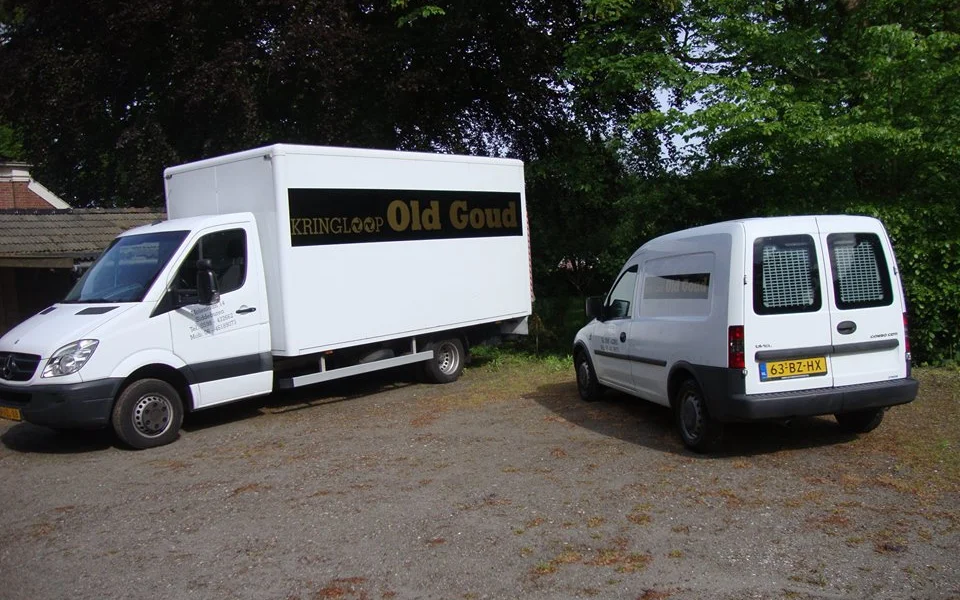 Twee witte bedrijfsbusjes van Kringloop Old Goud geparkeerd op een terrein met bomen op de achtergrond