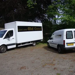 Twee witte bedrijfsbusjes van Kringloop Old Goud op een parkeerplaats tussen groen.