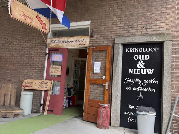 Ingang van kringloopwinkel met rode deur, houten deurkozijn en zwart bord met witte tekst aan de gevel