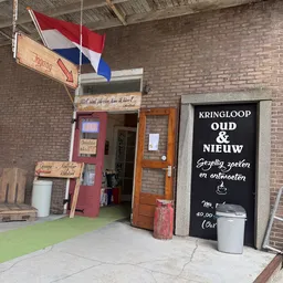 Ingang van kringloopwinkel met rode deur, houten deurkozijn en zwart bord met witte tekst aan de gevel