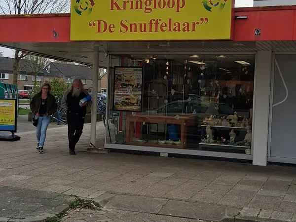 Gevel van kringloopwinkel De Snuffelaar met gele luifel en etalageruiten, twee personen lopen voorbij