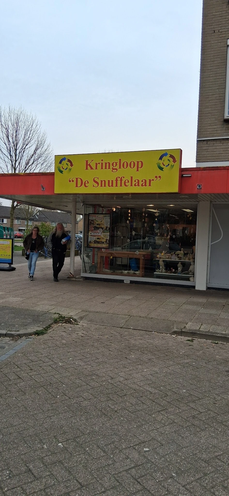 Gevel van kringloopwinkel De Snuffelaar met gele luifel en etalageruiten, twee personen lopen voorbij