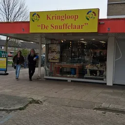 Gevel van kringloopwinkel De Snuffelaar met gele luifel en etalageruiten, twee personen lopen voorbij
