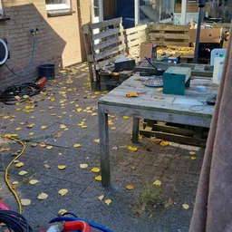 Buitenruimte/terras met werkbank en uitgestalde spullen, bladeren op de grond