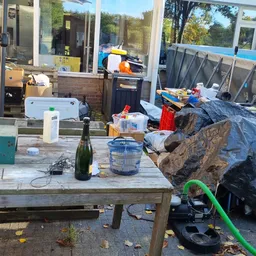 Rommelige buitenruimte met tafels, spullen en een grote steen, mogelijk achterplaats of berging van kringloopwinkel