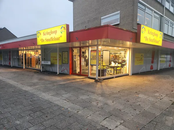 Gevel van kringloopwinkel met rode pui, gele letters en verlichte etalages aan het plein