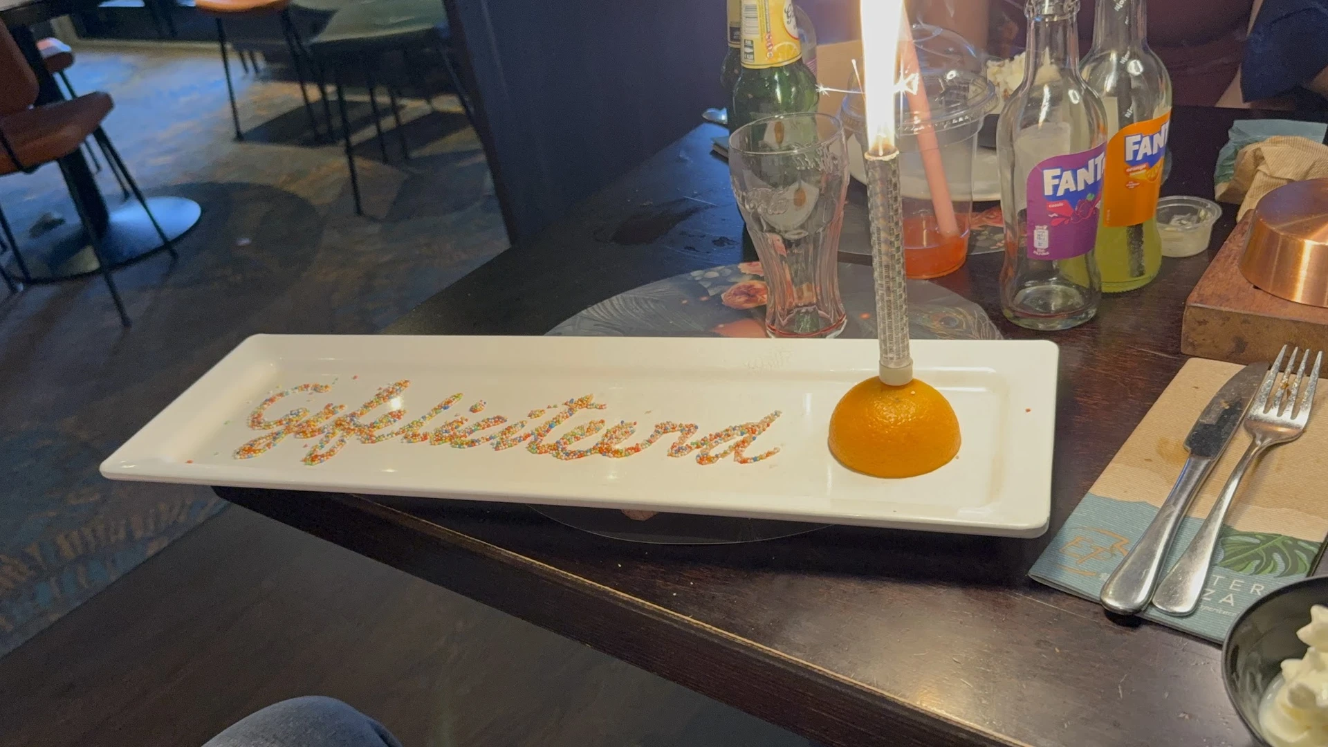 Dessertbord met tekst 'welkom' en een sinaasappel, glazen en flessen op achtergrond