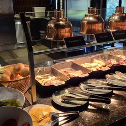 Warme dienbladen met eten onder koperen warmhoudkappen, bestek en serviesgoed op een buffettafel