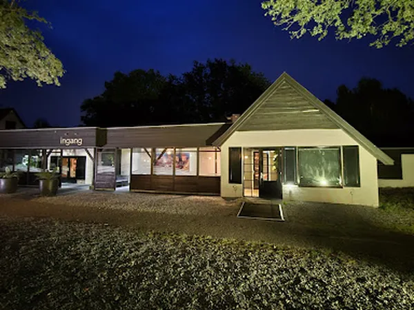 Avondfoto van een kringloopwinkel met verlichte pui en ingang, gelegen aan een grindduin