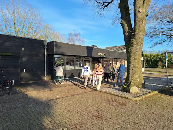 Zwart winkelgebouw met ingang, bezoekers buiten, grote boom naast pui
