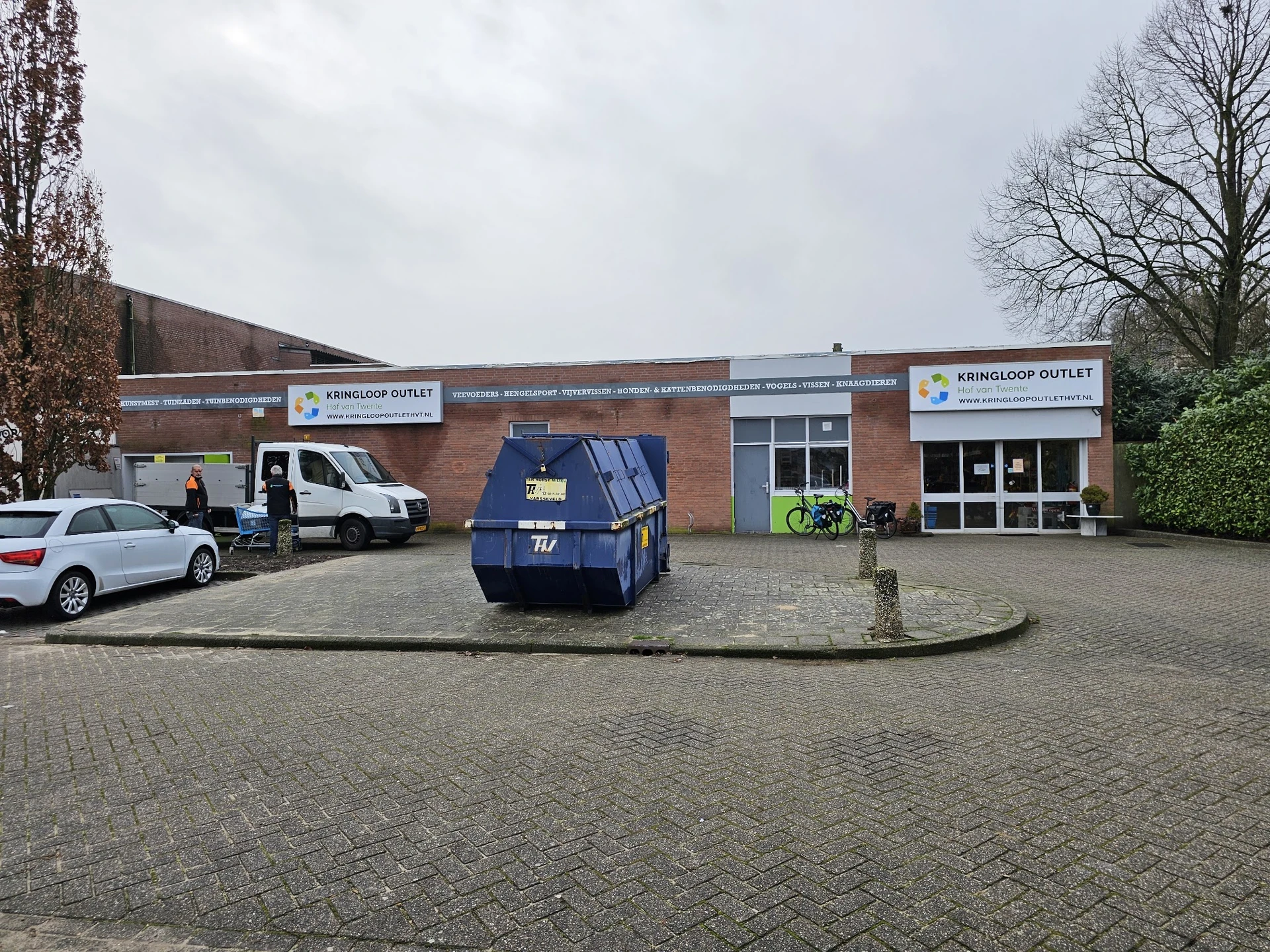 Voorgevel van bruine bakstenen kringloopwinkel met witte berding en twee ingangen, parkeerterrein met enkele auto's