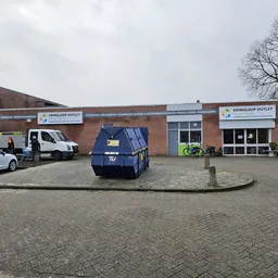 Voorgevel van bruine bakstenen kringloopwinkel met witte berding en twee ingangen, parkeerterrein met enkele auto's