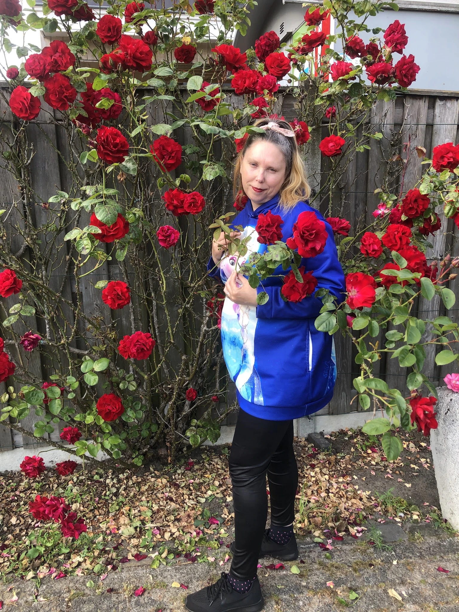 Vrouw in blauwe jas poseert met rode rozen tegen een met bloemen begroeide muur