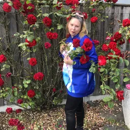 Vrouw in blauwe jas poseert met rode rozen tegen een met bloemen begroeide muur
