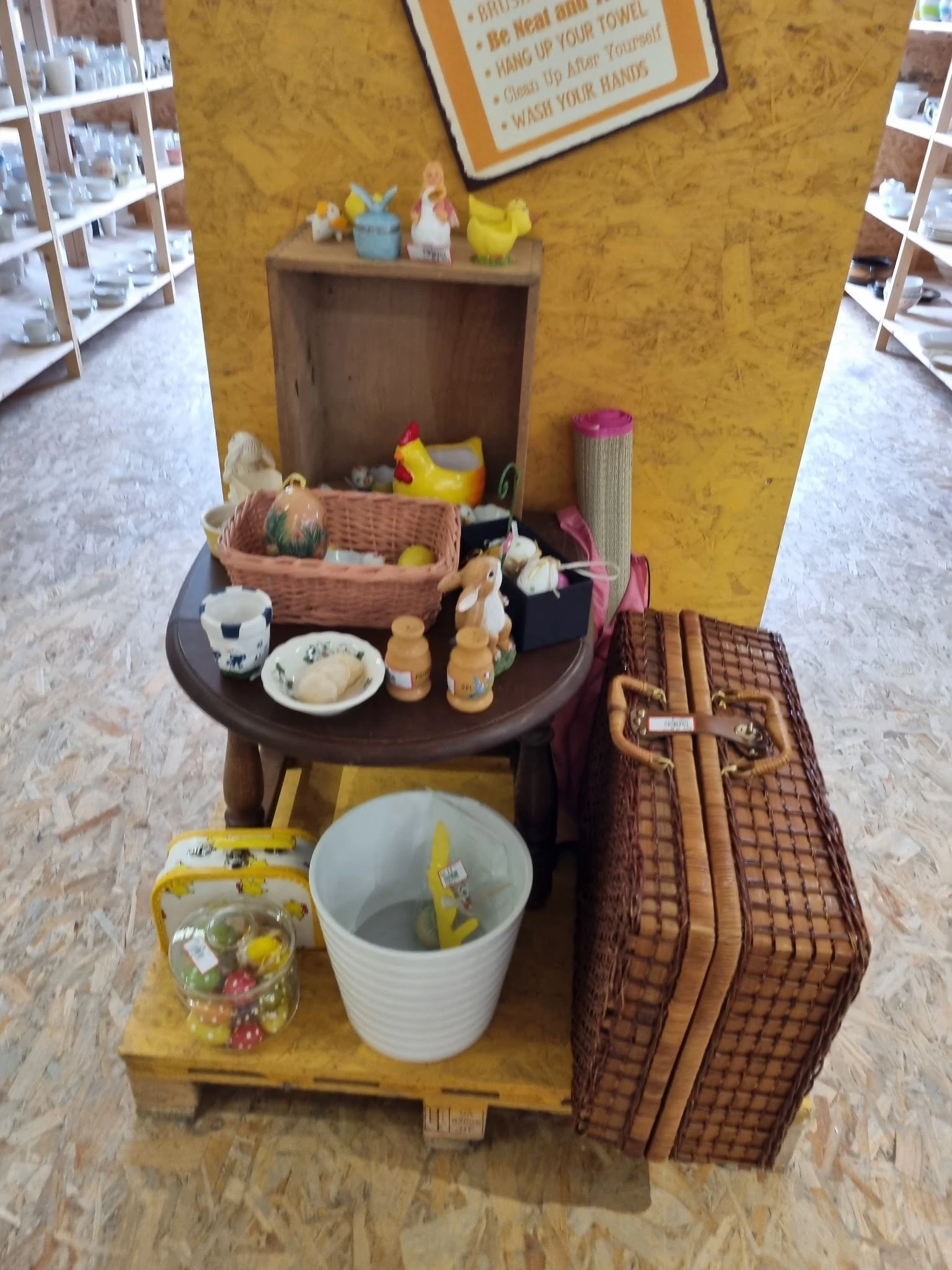 Uitgestalde tweedehands huishoudartikelen op tafel en plank, waaronder manden, servies en decoratieve items.