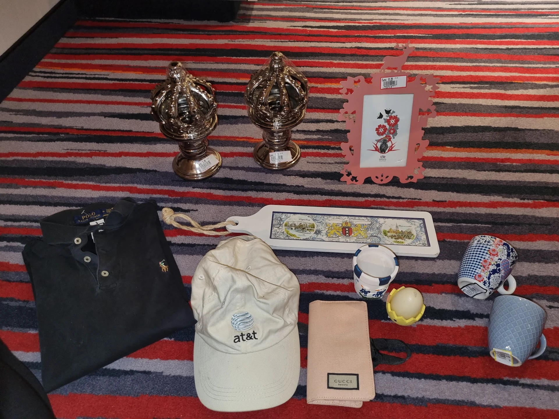 Uitgestalde tweedehandsartikelen op een gestreept tapijt: gouden beeldjes, poloshirt, at&t-pet, portemonnee, boeken en overig