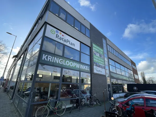Gevel van kringloopwinkel RataPlan met grote ramen, winkelnaam duidelijk zichtbaar, enkele fietsen ervoor, rode auto rechts